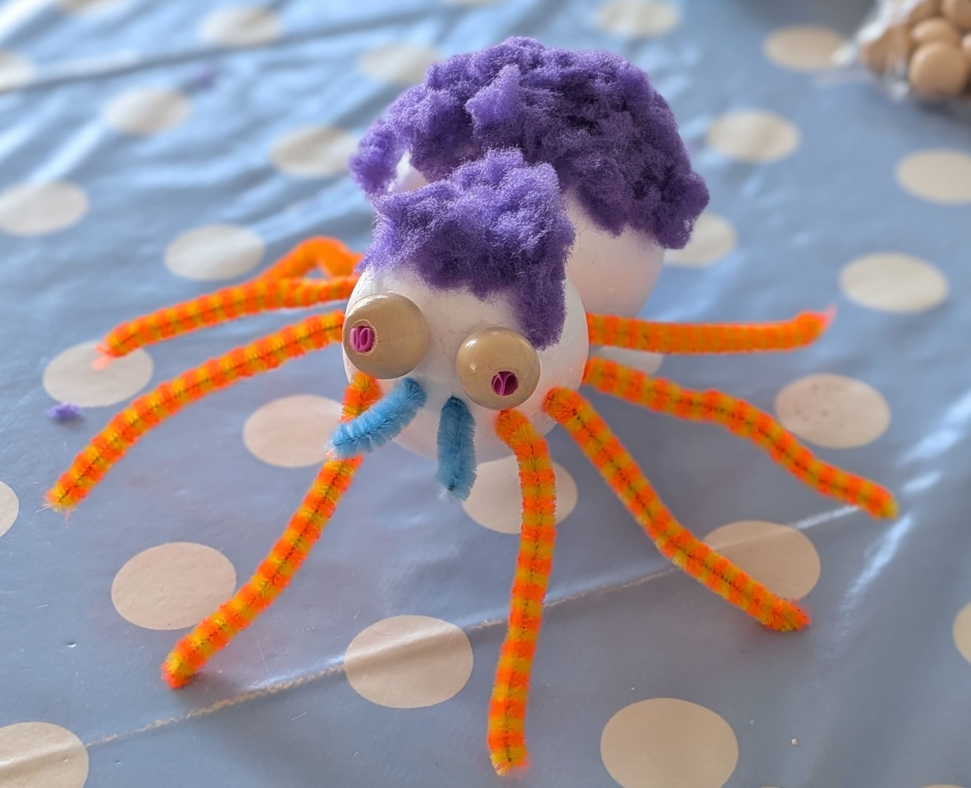 A homemade spider, with orange and yellow legs, blue tusks, big eyes with red pupils, and purple hair.