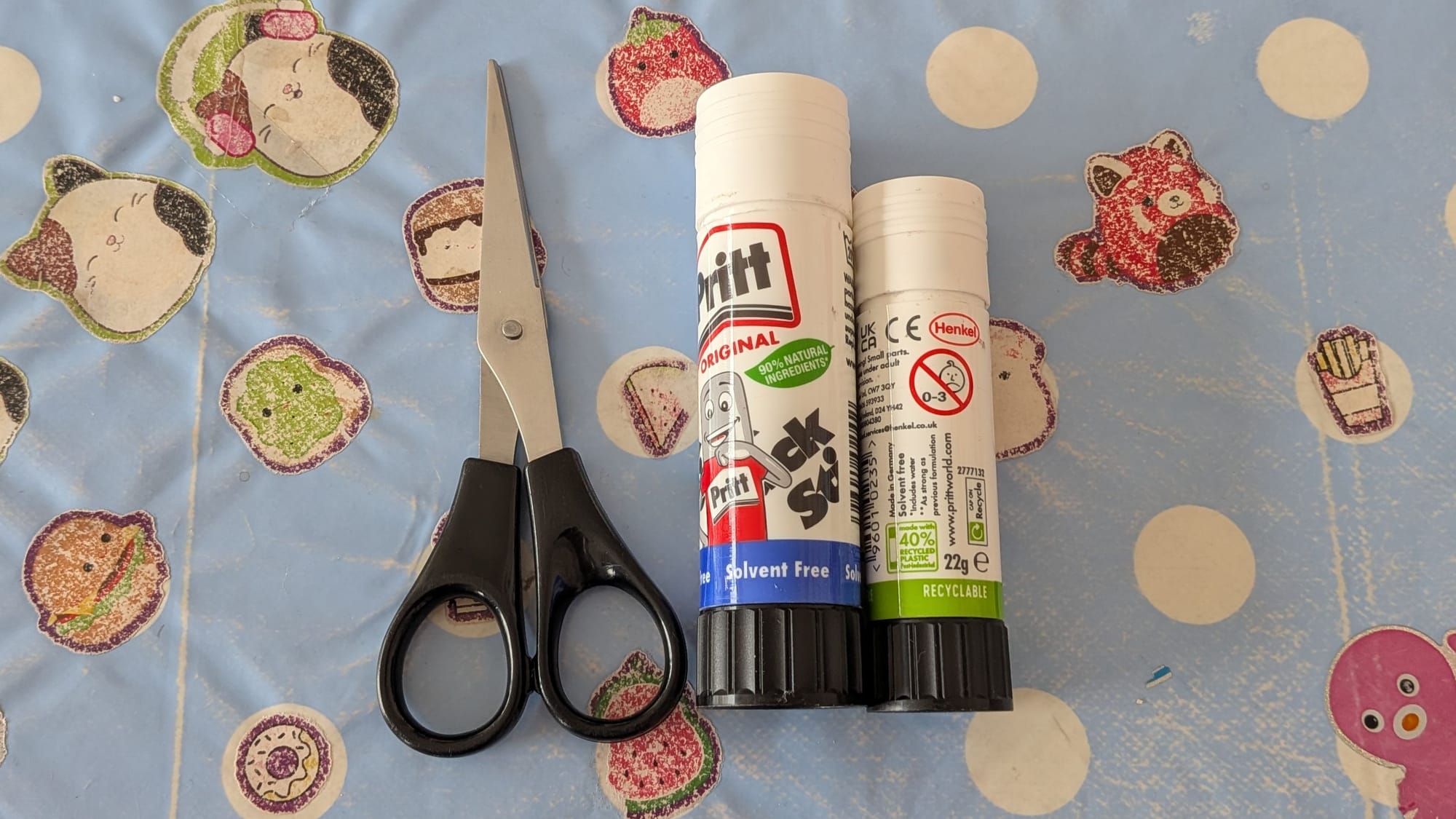 A pair of scissors (black handle) alongside two pritt stick (one small, one medium sized)