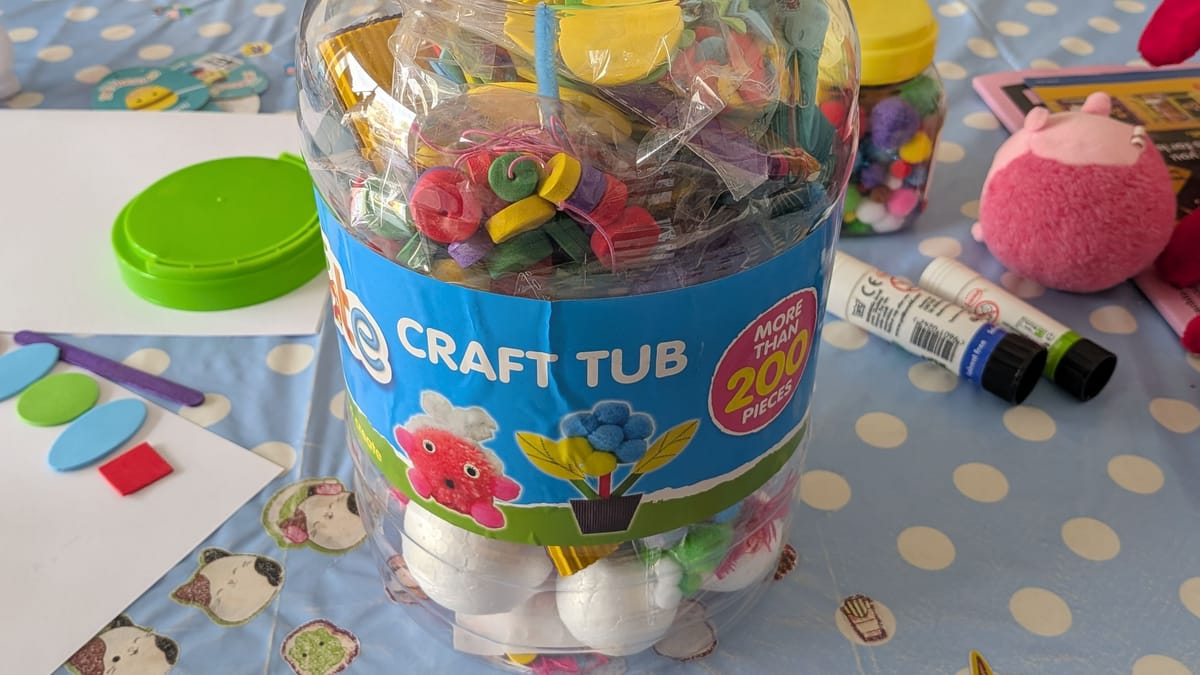 A plastic tub of crafting materials labelled "Craft Tub". To the side are glue sticks, paper, a pink soft toy, more crafting materials, and a green lid.