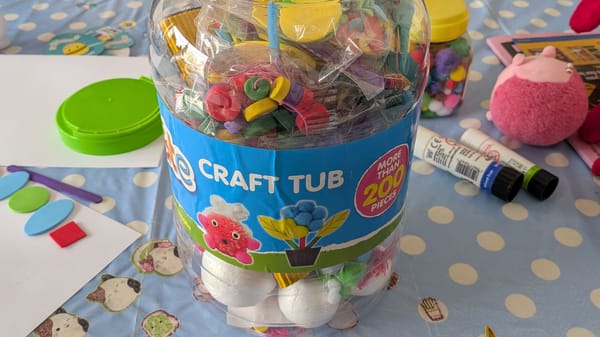 A plastic tub of crafting materials labelled "Craft Tub". To the side are glue sticks, paper, a pink soft toy, more crafting materials, and a green lid.
